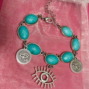 May include: Silver-toned bracelet featuring oval turquoise-colored stones and circular charms with an eye symbol. A dangling eye charm is also present. The bracelet is displayed on a pink surface.