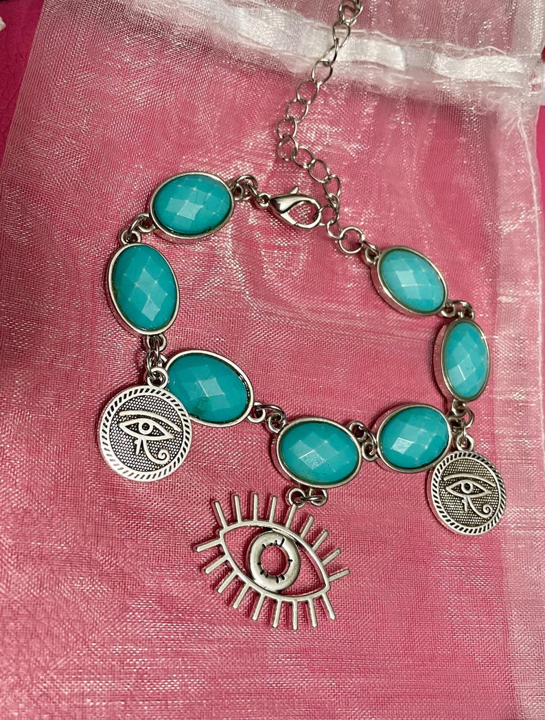 May include: Silver-toned bracelet featuring oval turquoise-colored stones and circular charms with an eye symbol. A dangling eye charm is also present. The bracelet is displayed on a pink surface.