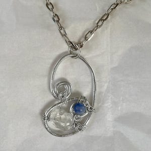 May include: Silver-toned pendant necklace with a clear crystal and a blue gemstone. The pendant has a wire-wrapped design with an oval shape and a chain link necklace.