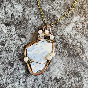 May include: A handcrafted necklace with a copper-coloured wire pendant holding a blue and white ceramic shard. The pendant is decorated with small white pearls and a clear crystal. The necklace has a gold-coloured chain.