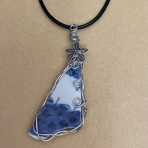 May include: A pendant necklace featuring a triangular piece of white ceramic with blue floral patterns, wrapped in silver-toned wire. The pendant is topped with a small silver-toned starfish charm and hangs from a simple black cord necklace.
