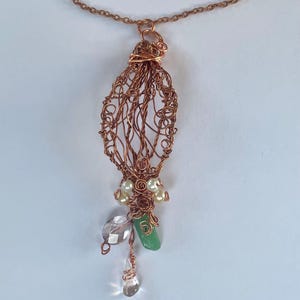 May include: A copper wire wrapped pendant necklace featuring an elongated cage design. The pendant is adorned with small cream-coloured pearls, a faceted clear crystal bead, a smooth green stone, and a small clear teardrop-shaped crystal bead.
