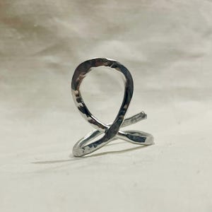 May include: A silver-colored, hammered metal ring with a unique, abstract design. The ring features a looped shape with intersecting lines, creating a modern and artistic look. The metal has a textured surface.
