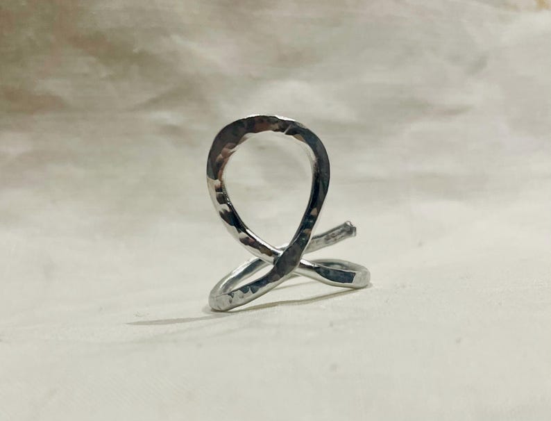 May include: A silver-colored, hammered metal ring with a unique, abstract design. The ring features a looped shape with intersecting lines, creating a modern and artistic look. The metal has a textured surface.