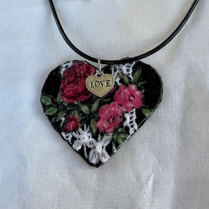 May include: Heart-shaped pendant necklace with a black background, adorned with red roses and green leaves. A small gold-colored charm with the word "LOVE" hangs from the top. The pendant is trimmed with white lace and hangs from a black cord.