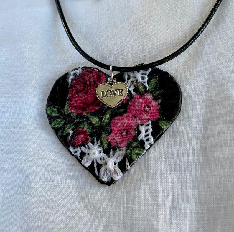 May include: Heart-shaped pendant necklace with a black background, adorned with red roses and green leaves. A small gold-colored charm with the word "LOVE" hangs from the top. The pendant is trimmed with white lace and hangs from a black cord.