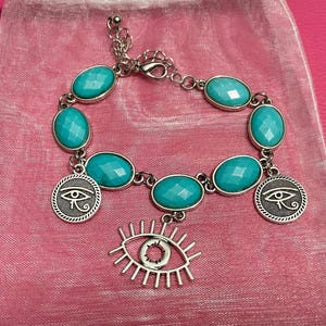 May include: Silver-toned bracelet featuring oval, faceted turquoise-colored stones. The bracelet includes two round charms with an eye design and a dangling eye charm. The bracelet is displayed on a pink surface.