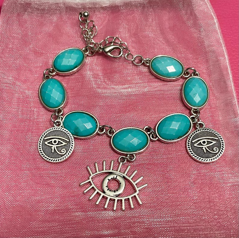 May include: Silver-toned bracelet featuring oval, faceted turquoise-colored stones. The bracelet includes two round charms with an eye design and a dangling eye charm. The bracelet is displayed on a pink surface.