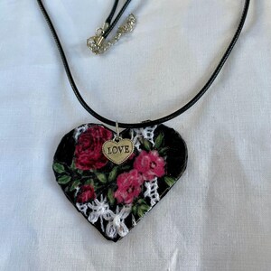 May include: A heart-shaped pendant necklace with a black background, adorned with red roses and white lace. A small gold heart charm with the word "LOVE" hangs from the pendant. The necklace has a black cord.