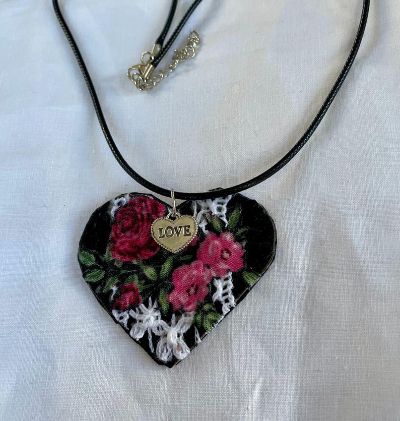 May include: A heart-shaped pendant necklace with a black background, adorned with red roses and white lace. A small gold heart charm with the word "LOVE" hangs from the pendant. The necklace has a black cord.