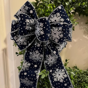 May include: A large navy blue bow with silver snowflake patterns and a silver trim. The bow is set against a backdrop of green foliage, creating a festive, wintery aesthetic. This decorative bow is suitable for holiday decor.