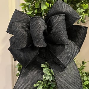 May include: A close-up of a decorative wreath featuring a large, textured black bow. The bow is prominently displayed against a backdrop of lush, green foliage. The bow's ribbon has a woven appearance, adding depth and texture to the design.