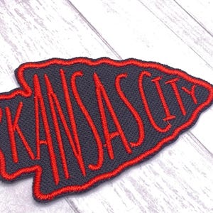 May include: Embroidered patch in the shape of an arrowhead, featuring the text "KANSAS CITY" in red stitching on a dark gray background. The patch has a red border and is likely for apparel or accessories.