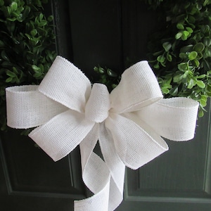 May include: A white burlap bow with a long tail, tied to a green wreath.