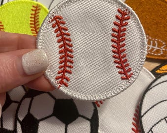 May include: A white baseball patch with red stitching details. The patch is round and features the classic baseball design. Other sports patches, including soccer balls, softballs, and volleyballs, are visible in the background.
