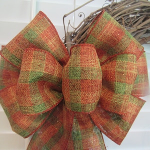 May include: A large decorative bow with a plaid pattern in shades of orange, green, and gold. The bow is made of a textured fabric and is likely used for seasonal decor or gift wrapping. The bow is centered in the frame.