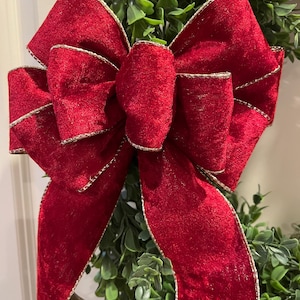 May include: A close-up of a festive wreath featuring a large, plush red velvet bow with gold trim. The bow is the focal point, set against a backdrop of vibrant green foliage. The bow's texture and color create a striking visual contrast.