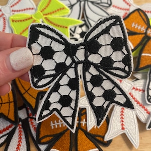 May include: A collection of embroidered bow patches in various sports themes. The central bow features a soccer ball design with black and white detailing. Other bows include baseball, football, and a solid neon yellow bow.