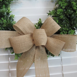 May include: A burlap bow tied around a green boxwood wreath. The wreath is hanging on a white slatted blind.