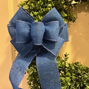 May include: A blue burlap bow with long tails is attached to a green wreath. The bow is made of a coarse, woven material and has a layered design. The wreath is made of small, green leaves.
