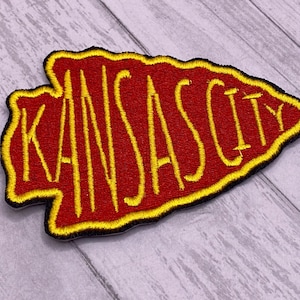 May include: Embroidered patch in the shape of an arrowhead, featuring the text "KANSAS CITY" in yellow thread on a red background. The patch has a yellow border and a black outline.