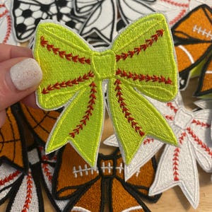 May include: A collection of embroidered bow patches in various sports themes. The central bow is lime green with red stitching, resembling a softball. Other bows feature baseball, football, and soccer designs, with white, orange, and black accents. These patches are ideal for crafting and personalization.