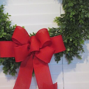 May include: A green wreath with a large red bow. The wreath is made of artificial greenery and the bow is made of a soft, textured fabric.