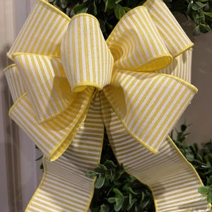 May include: A yellow and white striped bow with a wired edge. The bow is tied around a green bush.