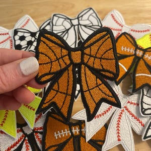 May include: Embroidered patches in the shape of bows, featuring sports themes. The patches include designs for basketball, baseball, and football. The basketball bow is orange with black detailing. Other bows are white, orange, and green.