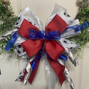 May include: A decorative bow with red, white, and blue ribbons, featuring soccer ball patterns. The bow is part of a wreath with greenery and wooden beads, suitable for sports-themed decor.