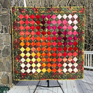 May include: A square quilt featuring a gradient of diamond-shaped fabric patches transitioning from yellow and orange to red, pink, and white. The quilt is framed by a dark green border with a floral pattern and a thin yellow binding edge.