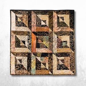 May include: A square quilt featuring a geometric block pattern composed of various fabric squares in shades of black, tan, cream, and burnt orange. The design includes intricate batik-style prints, floral motifs, and swirling patterns throughout.