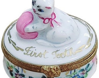 May include: A round white porcelain trinket box with a gold-tone metal hinge. The lid features a sculpted white cat with a pink bow playing with a pink ball, and the text First Tooth in gold script. The base is decorated with painted pink and blue flowers.