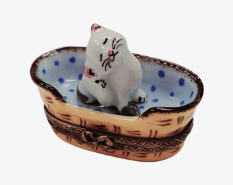May include: A porcelain trinket box shaped like a basket with a light blue interior featuring dark blue polka dots. A small white cat figurine with black markings and pink ears sits inside the basket. The exterior has a tan finish with brown markings.