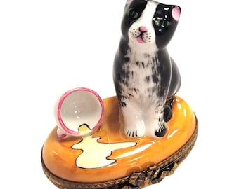 May include: A porcelain trinket box featuring a black and white cat sitting on an oval orange base. Next to the cat is a small overturned white cup with a pink rim and a spilled puddle of white liquid. The base has a decorative metal hinge.
