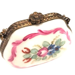 May include: A small, oval-shaped porcelain trinket box with a white base, featuring a hand-painted floral motif in pink, blue, and green. The container has a metallic, bronze-toned frame with a flower-shaped clasp and a small attached chain.