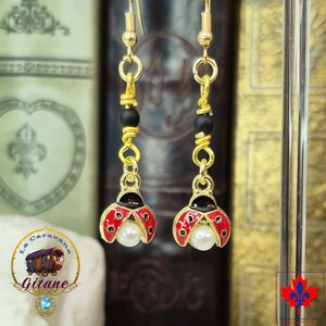 May include: Gold-coloured ladybird earrings with red enamel wings and black detailing. Each earring has a faux pearl and black bead accents. The earrings are suspended from gold-coloured ear wires.