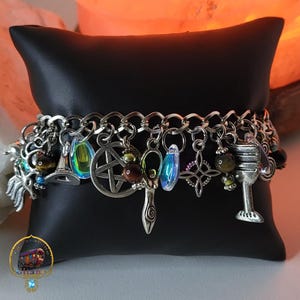 Puede incluir: Un brazalete de plata con una variedad de dijes, incluyendo un pentagrama, un sombrero de bruja, una copa y un cristal. El brazalete está sobre un fondo de terciopelo negro.