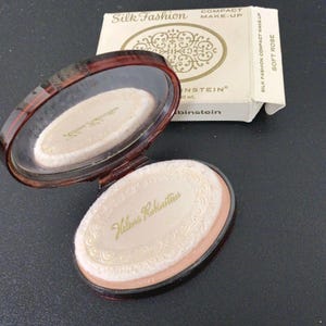 May include: An oval Helena Rubinstein compact make-up case in a dark brown translucent finish with an open lid revealing a mirror and a powder puff. The accompanying white cardboard box is labelled Silk Fashion Compact Make-Up Soft Rose.