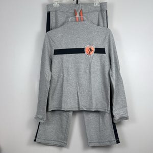 Puede incluir: Conjunto de chándal gris, que incluye una chaqueta con cremallera y pantalones a juego. La chaqueta presenta una franja negra en el pecho con un logotipo y un cuello alto. Los pantalones tienen una franja negra en el lateral.