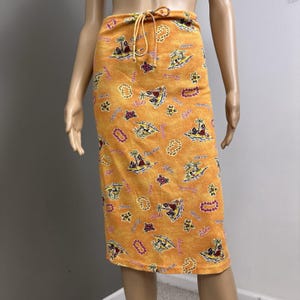 May include: Orange knee-length skirt with a tropical print. The skirt features a drawstring waist and is covered in images of palm trees, islands, and the word "Aloha". The skirt is made of a soft, lightweight fabric.