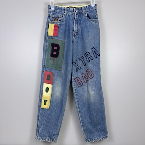 Pode incluir: Jeans azuis com patches coloridos e texto bordado. Os patches incluem um design de blocos de cores vermelho, amarelo e verde e um patch verde com a letra "B" e as palavras "BUBLE BOY". O texto "XTRA BAD" é bordado na perna direita.