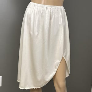 May include: A white satin half slip with an elastic waistband and a slit on the side. The slip has a scalloped hemline. The garment is displayed on a mannequin.