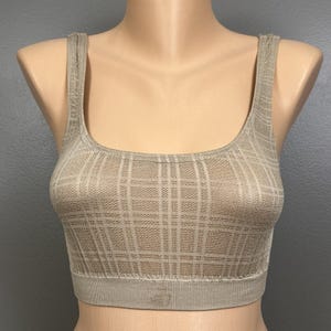 May include: A beige crop top featuring a square neckline and wide shoulder straps. The garment is constructed with a textured, knit fabric displaying a subtle plaid pattern throughout. A ribbed hem band sits at the base of the top.