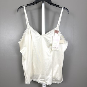 Puede incluir: Camisola de color marfil con tirantes ajustables y escote en V. La camisola tiene un delicado ribete de encaje a lo largo del escote y el dobladillo. Una etiqueta con la marca "HONORS" está adjunta. La prenda está colgada de una percha negra.