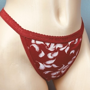 May include: Red thong underwear with a white feather pattern. The waistband features the text "VICTORIA'S SECRET" in white. The edges are trimmed with a scalloped lace detail.