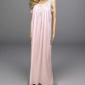 May include: A floor-length light pink nightgown featuring a scoop neckline with cream-colored lace trim, a small central bow, and two buttons. The garment has short, ruffled lace sleeves and a flowing, loose-fitting silhouette made of sheer fabric.