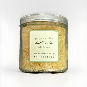 May include: A clear glass jar filled with yellow bath salts, featuring a silver screw-top lid and a pale green label. The label reads: paperwhite bath salts, sels de bain, net wt. 16 oz / 453 g, POTTERY BARN. The contents are granular.