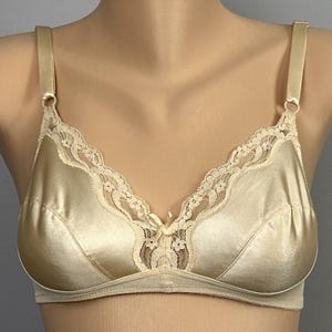 May include: A beige satin bra with lace trim along the neckline and a small bow in the center. The bra has adjustable straps and a classic design, offering a smooth, comfortable fit. The bra is displayed on a mannequin.