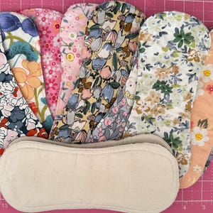 May include: A collection of seven reusable cloth pads featuring various floral and botanical patterns in shades of blue, pink, yellow, green, and peach, arranged in a fan shape behind a single plain, unbleached cream-colored cloth pad.
